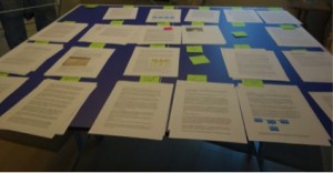 A blue table covered with numerous sheets of printed documents and several green sticky notes arranged in groups.